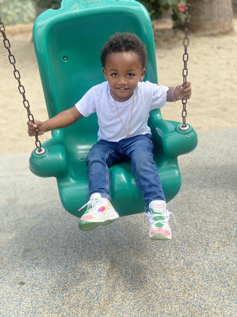 A toddler boy is happy swinging on a green swing at the playground.