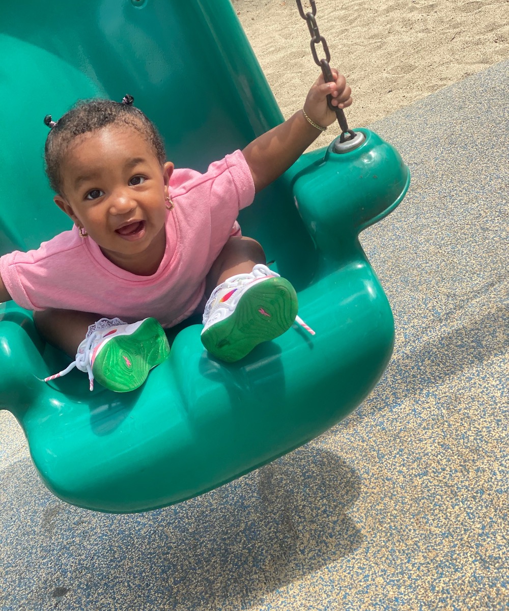 Toddler girl in a pink dress on a green swing at the park smiling for a picture.